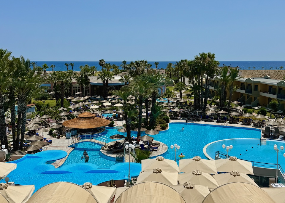 A large pool with attached jacuzzis and kids pool set in grounds filled with palm trees and facing the sea at Atrium Palace resort in Lindos.