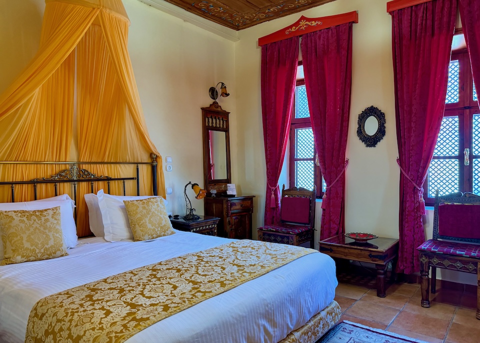 Vibrant yellow and red brocade bedding, curtains, and a canopy in a guest room with a hand-painted ceiling and terra cotta floor at Nikos Takis Hotel in Rhodes Old Town.