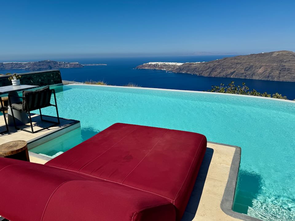 View across a red sun bed over an infinity pool with views of the Santorini Caldera and cliff towns in the distance