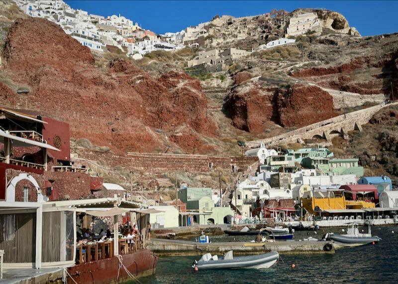 Boats and waterfront restaurants lining Ammoudi Bay. The staircase down from Oia village is visible just behind.