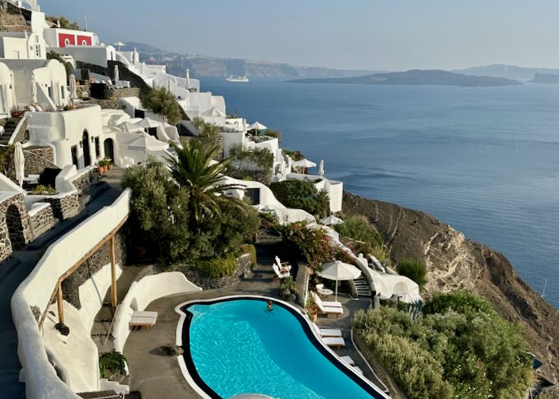 View over a white hotel with blue swimming pool, spilling down the Santorini cliffside.