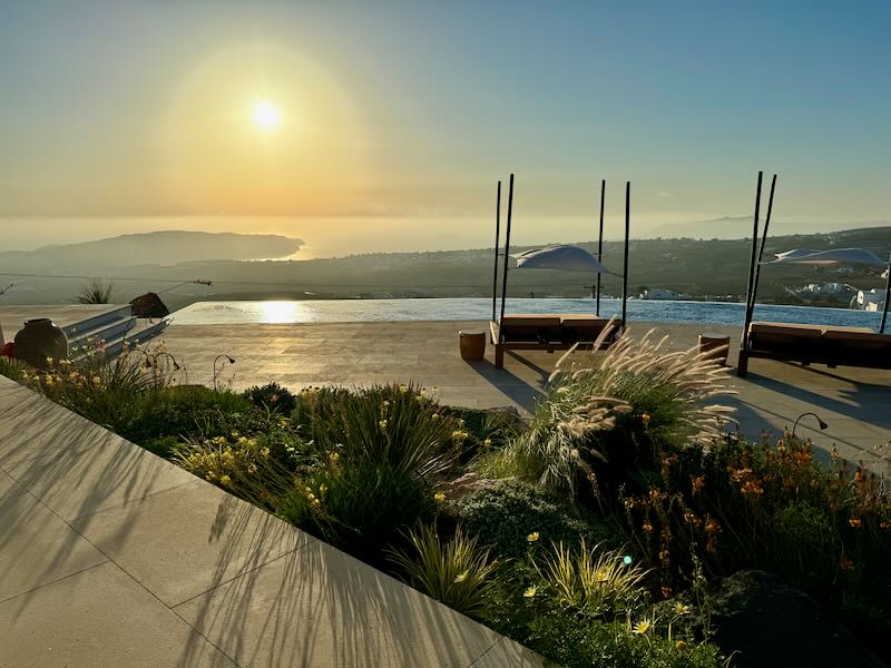 View of the sunset from an infinity pool deck with two shaded sun beds and mediterranean landscaping.