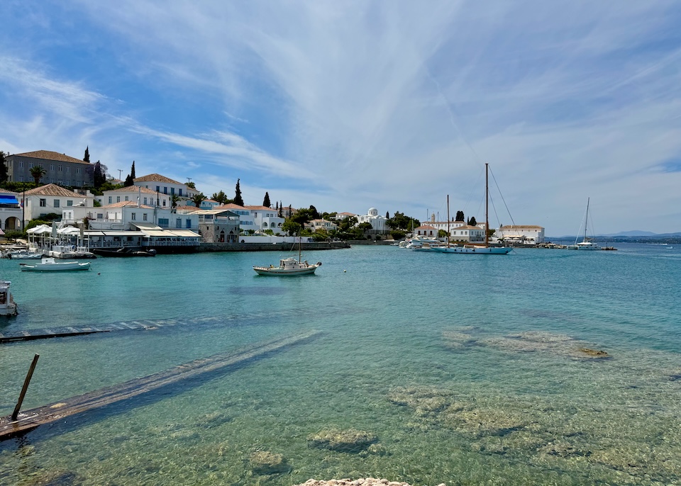 View of a harbor with clear water, boats, and a small town with red tile roof buildings in the Old Harbor of Spetses.