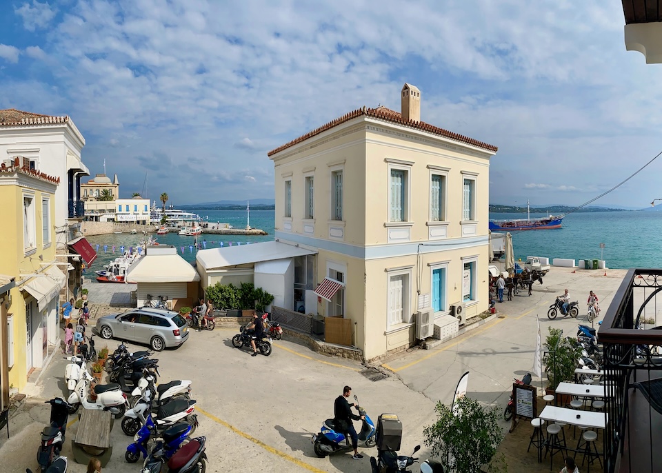 A busy little seafront road with a neoclassical building in the middle, the horse and carriage station, and scooters parked and driving in Dapia.