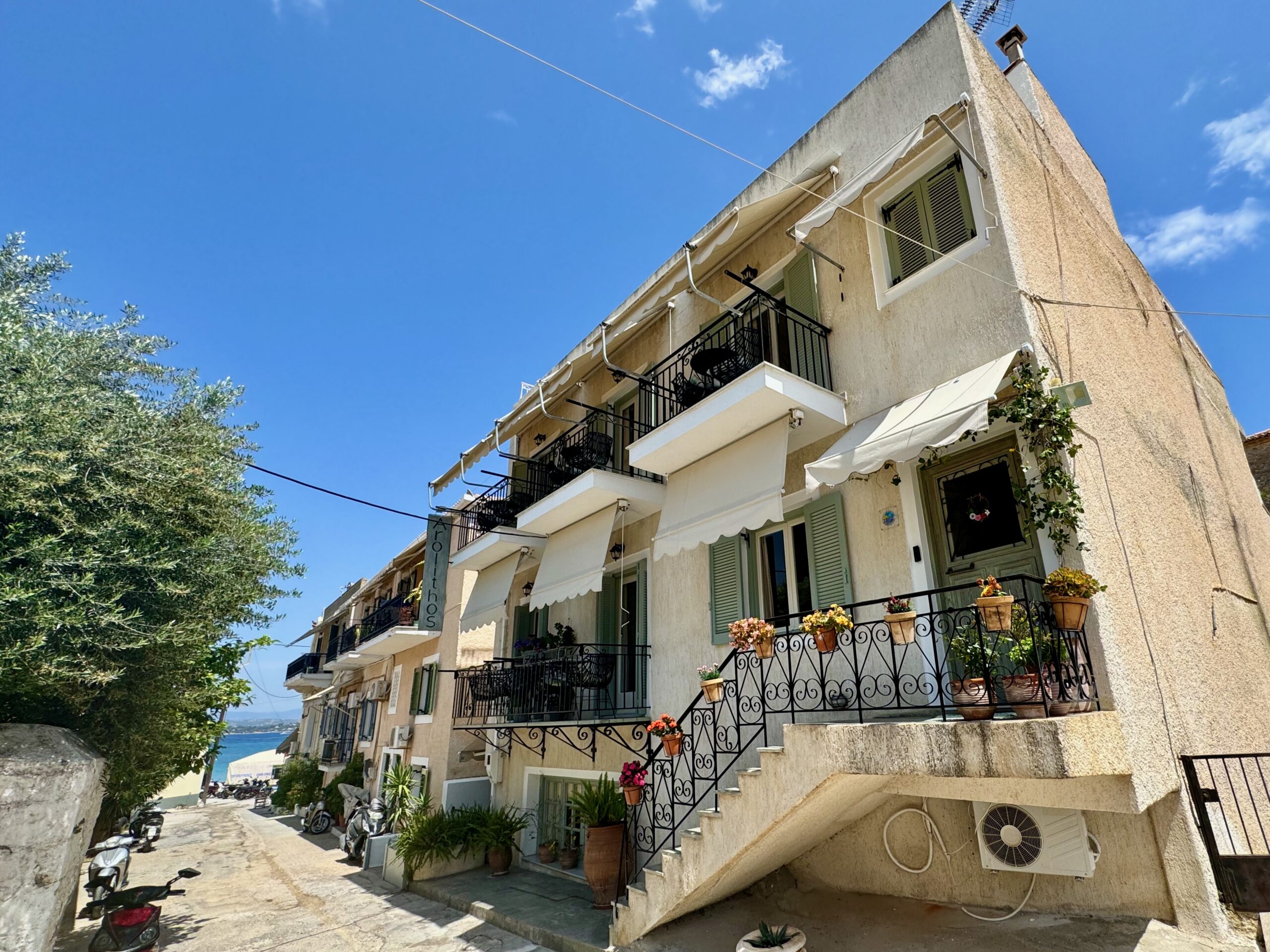 Exterior view of Arolithos hotel with two stories of balconies, potted plants, and the sea in the background in Spetses.