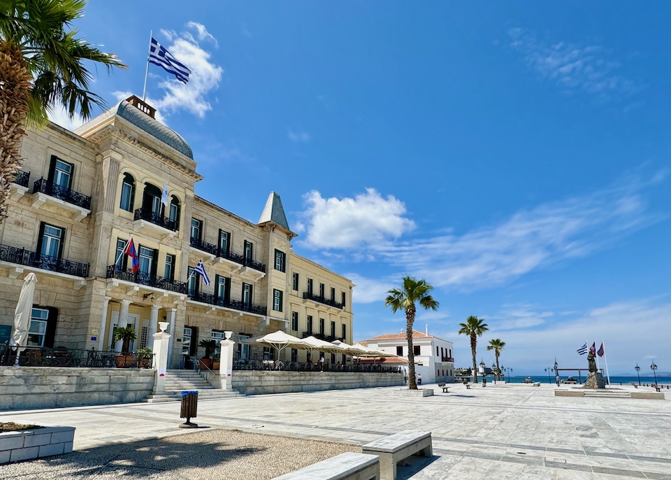 An imposing neoclassical building with three stories on a marble-paved square with the sea in the background.