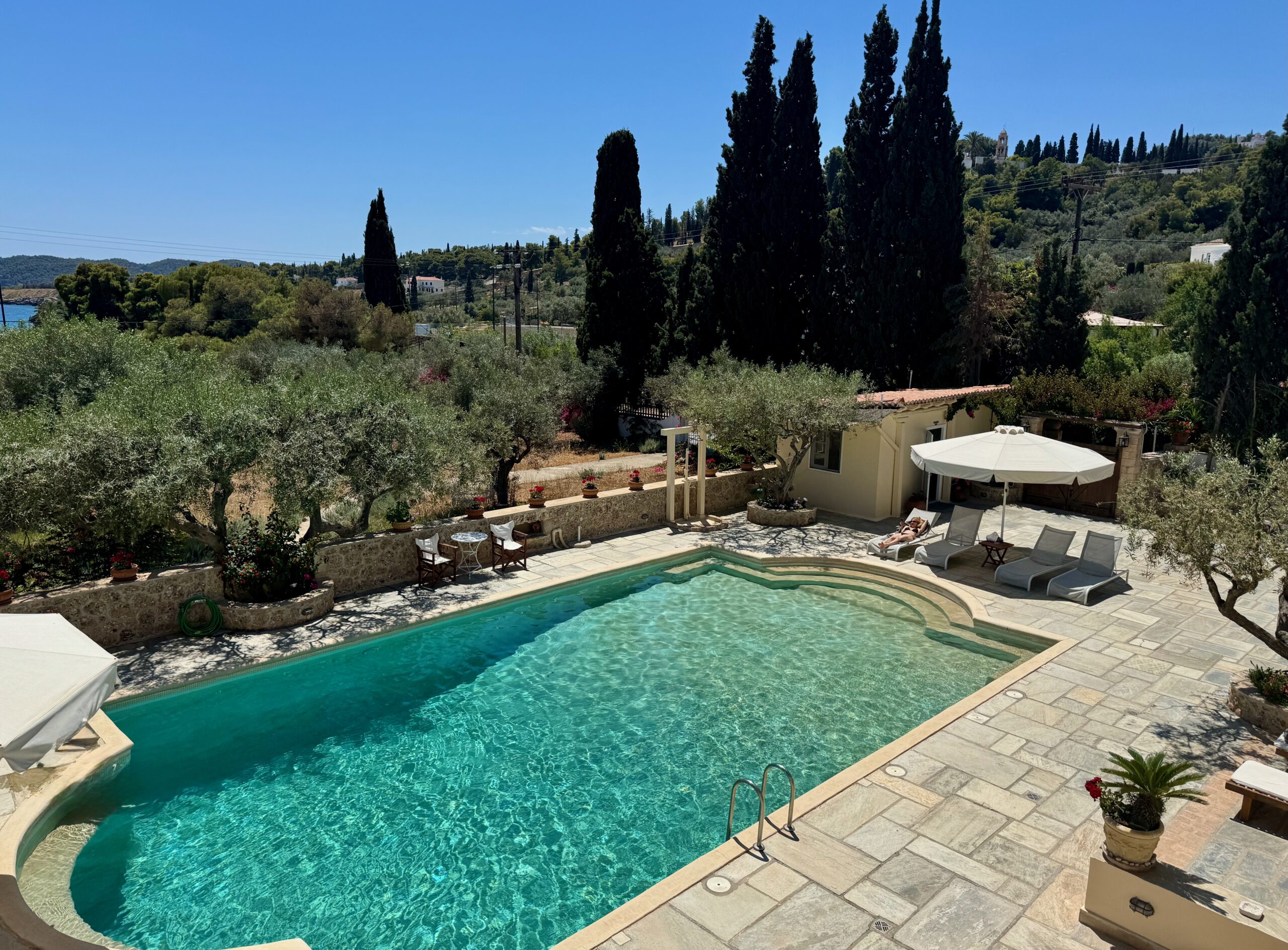 View from above a dazzling pool with sunbeds and one end, olive and cypress trees and the sea in the distance at Villa Nika in Spetses.