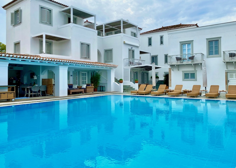 A pool with a row of sunbeds on one side and an restaurant on the other at Zoe's Club hotel in Spetses.