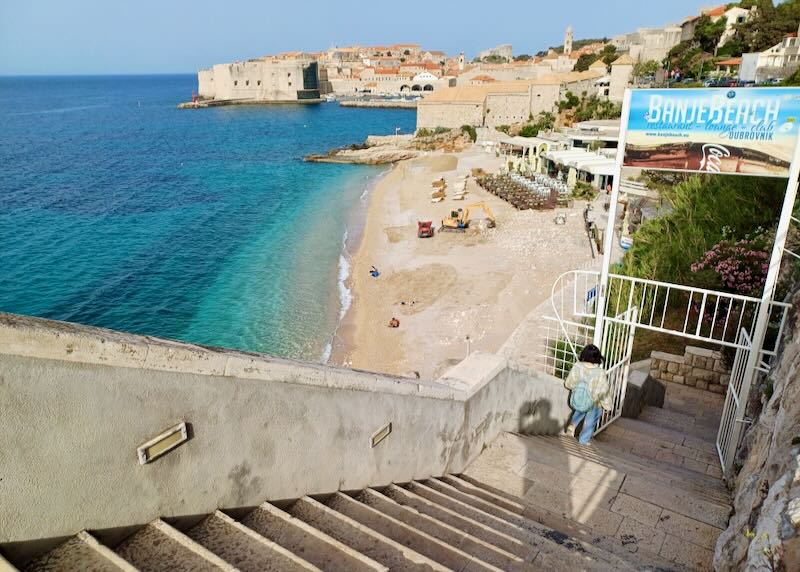 Outdoor stairs descend to a a beach with clear blue water.