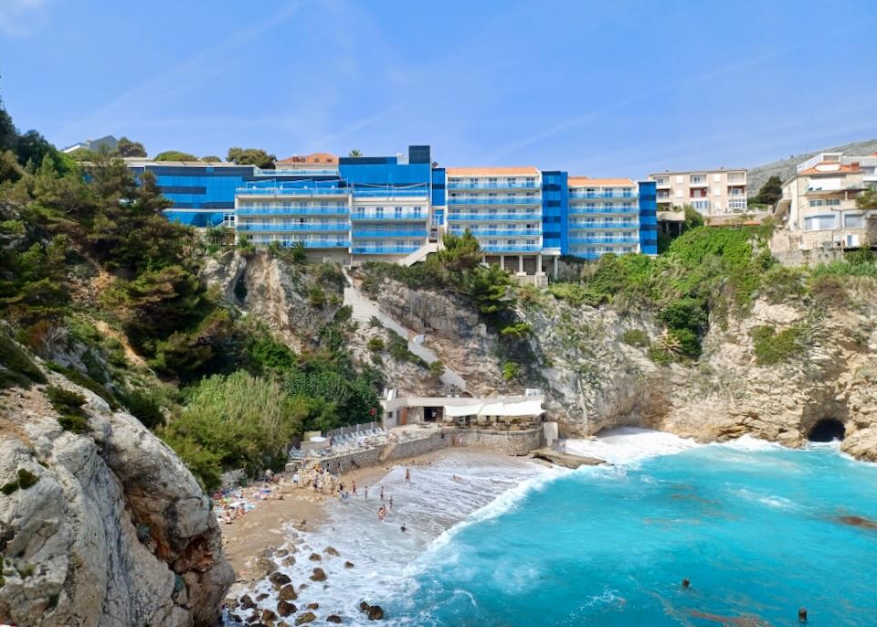 A hotel with sections of blue glass sits a top a cliff where below sits a beach.