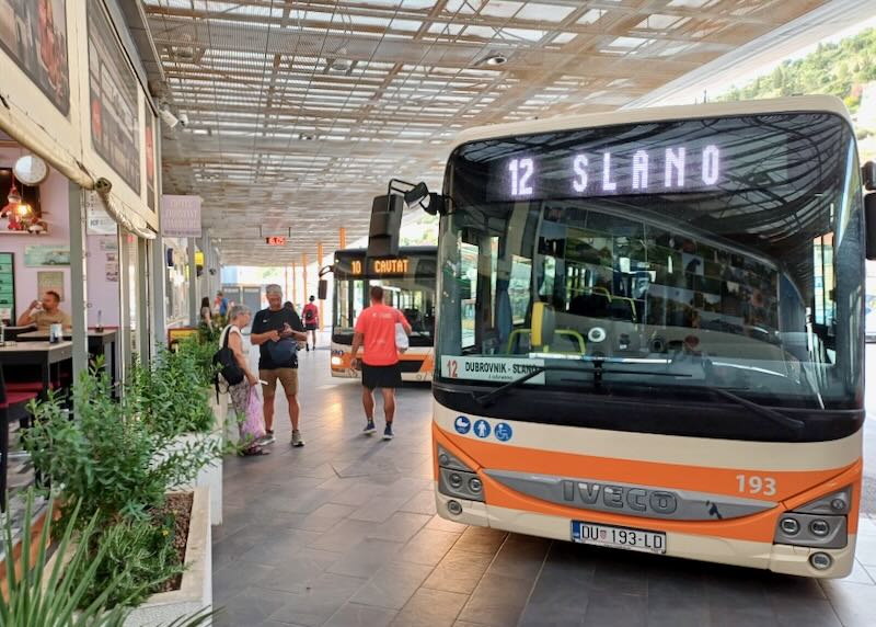 Travelers wait under a roof where several buses park and wait for riders.