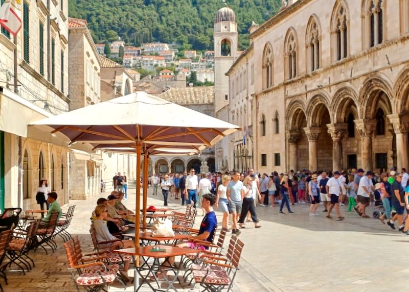 People walk through an old town while guests dine at outdoor tables.