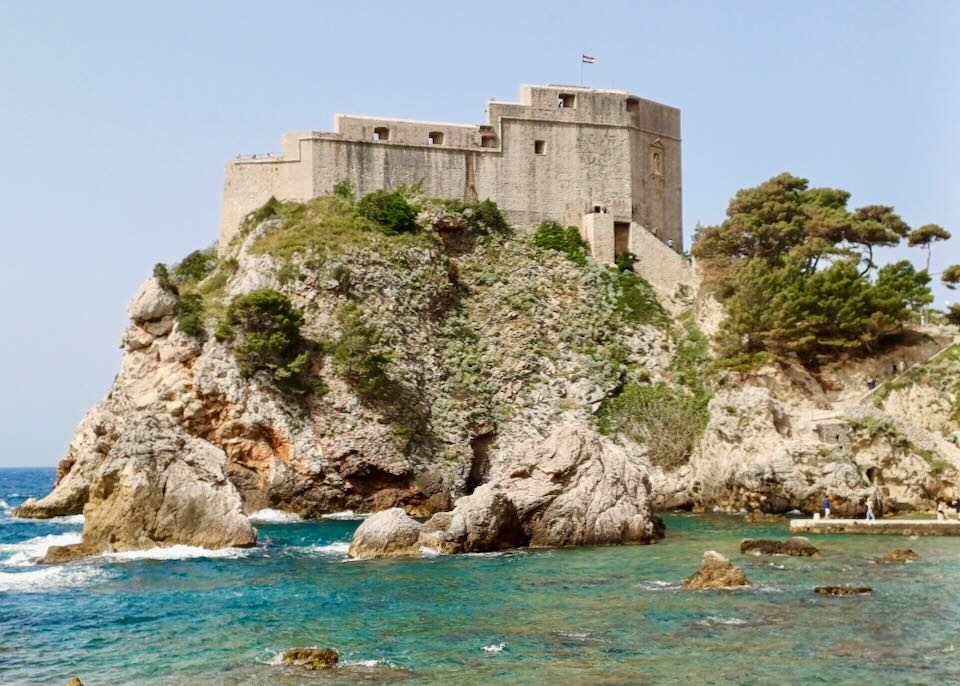 A tan stone historic fort sits on top of a tall gnarly rock with green bushes sprouting on it's side.