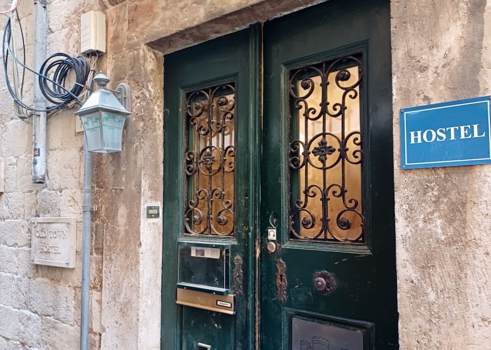 A blue sign that reads Hostel hangs by a green door with a curly wrought iron design on the windows.