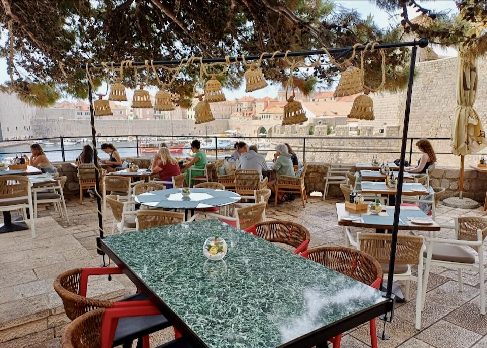 People sit at tables on a terrace eating meals and overlooking a port and old town.