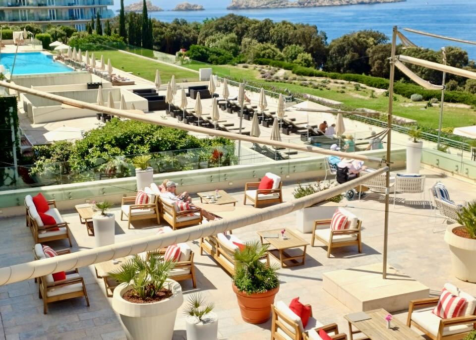 People sit at table and chairs, lounge chairs and the pool at a resort facing the sea.