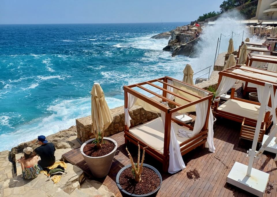 A couple sit next to daybeds and watch the sea crash into an ancient stone wall.