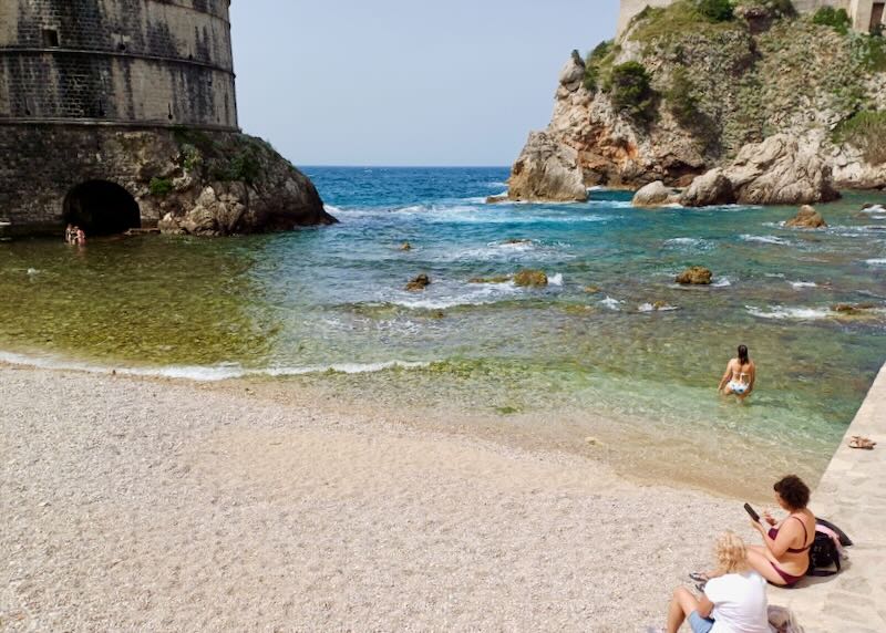 People wade into clear ocean water next to cliffs.