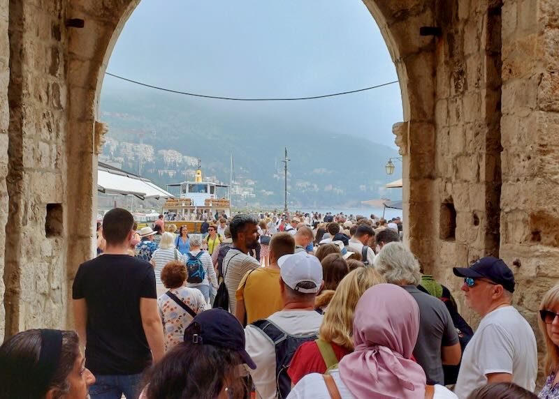 Crowds of people funnel through an opening in a stone wall to a port.