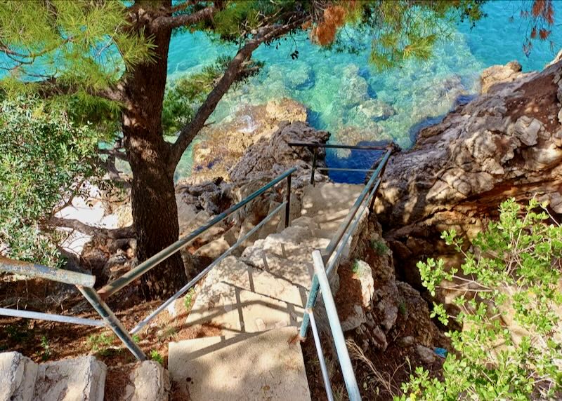 Steep outside stairs descend past a tree to clear blue water, exposing the rocks below.