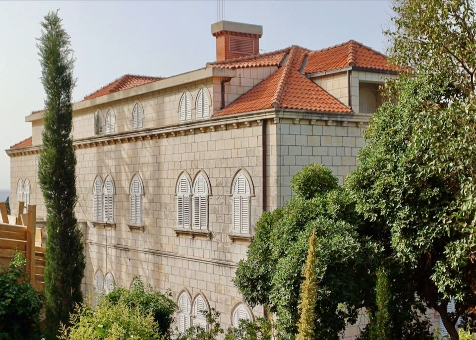 A villa with tan stone and arched white shutters and a red tiled roof sits on a hill.