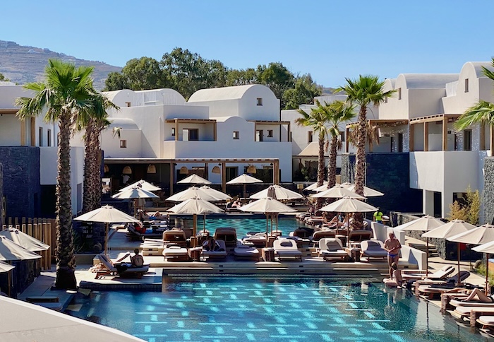 Two pools divided by a terrace with rows of sunbeds and umbrellas on it with palm trees and Cycladic buildings in the background at Radisson Blu Zaffron Resort in Santorini.