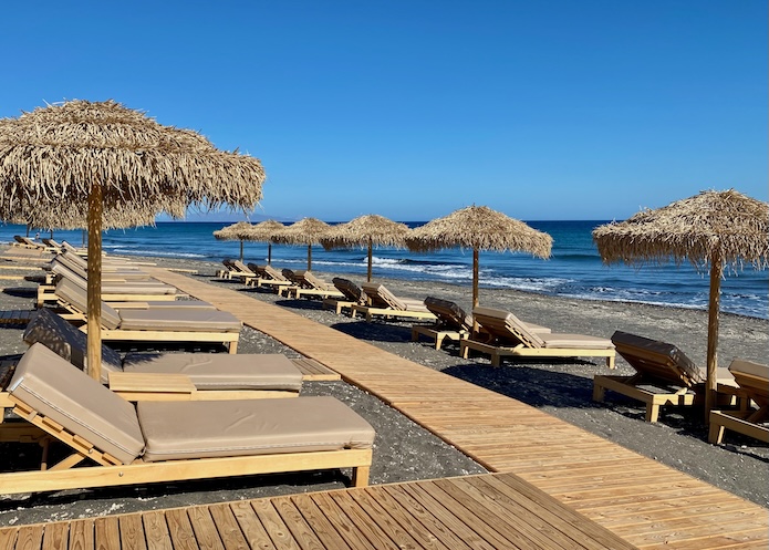 Pairs of sunbeds under thatched umbrellas line the beach, connected by a boardwalk at Sea Breeze Beach Resort in Santorini.