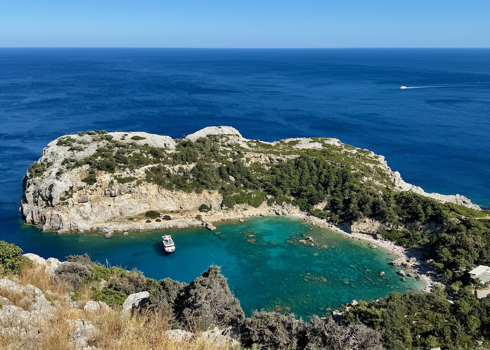 View over horseshoe-shaped Anthony Quinn Bay in Rhodes with vivid blue water and a boat moored inside.