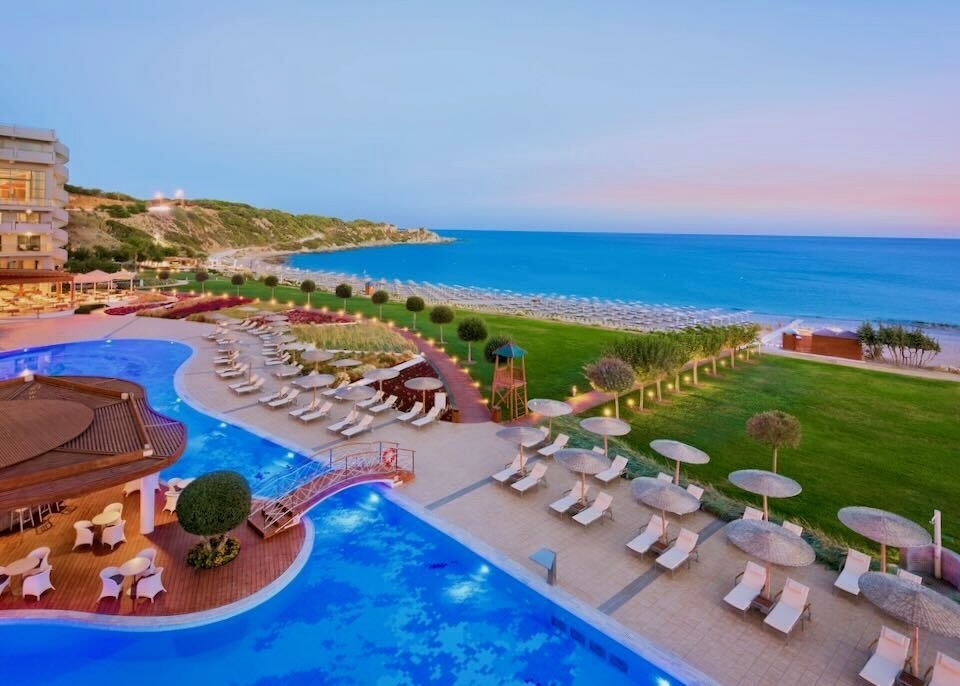 View over a pool with sunbeds and umbrellas and toward the beach at sunset at Elysium Resort in Rhodes.