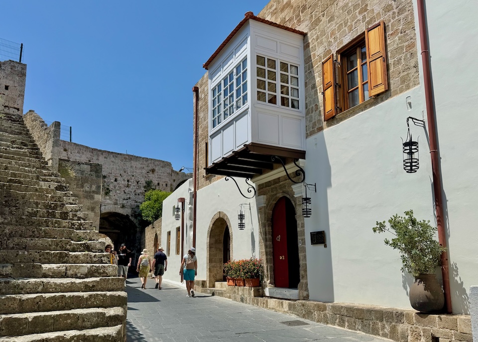 Exterior view of Kokkini Porta Rossa hotel in Rhodes Old Town with an Ottoman covered balcony above the street and stairs leading up to the medieval city walls across the road.