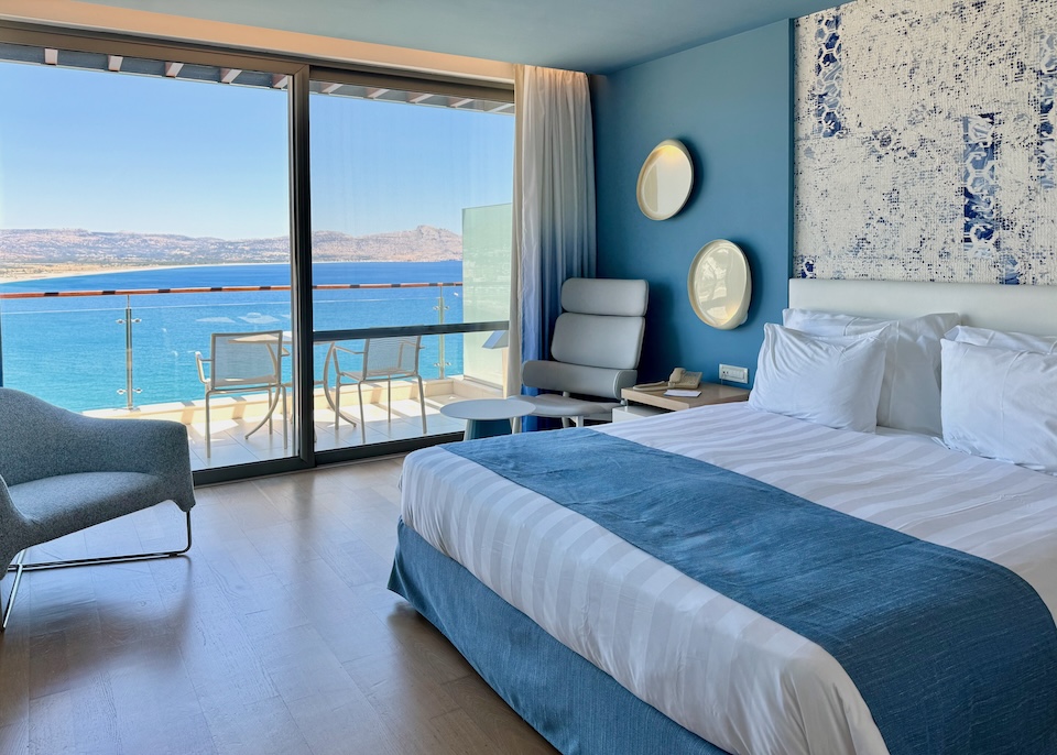 A contemporary hotel room in clean lines with blue and white colors and a sliding glass door leading out to a sea-view balcony at Lindos Blu resort in Rhodes.