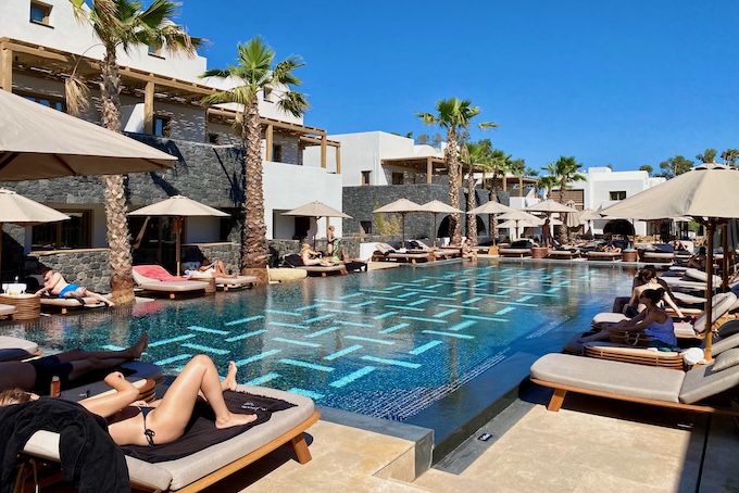 A rectangular, mosaic-tiled pool with a geometric design and sunbed-umbrella sets on all sides at Radisson Blu Zaffron Resort in Santorini.