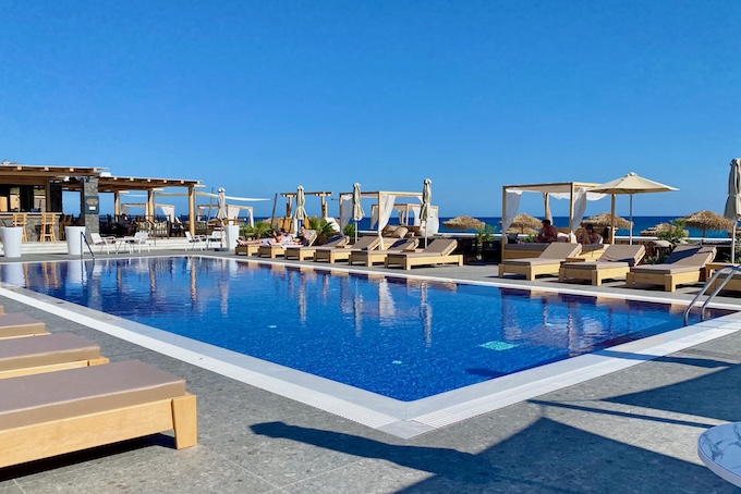 An infinity edge pool lined with sunbeds, Bali beds, and umbrellas with a pool bar at the far end and a view of the Aegean at Sea Breeze Beach Resort in Santorini.