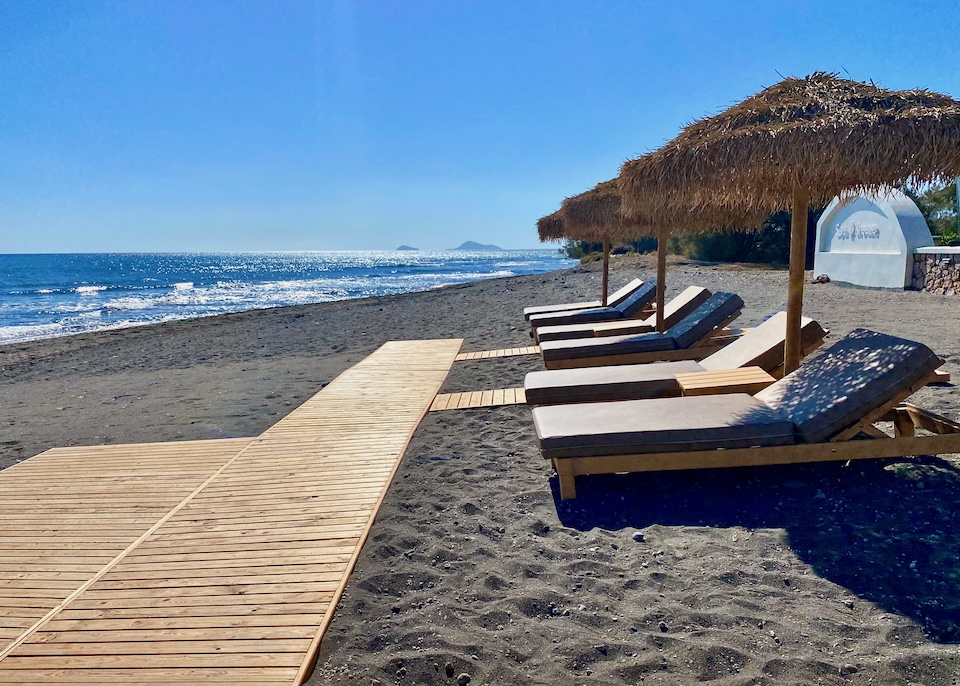 Pairs of sunbeds and thatched umbrellas sit on a black sand beach at the end of a wooden boardwalk at Sea Breeze Beach Resort in Exomitis, Santorini.