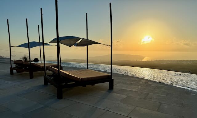 Two sun beds with canopies blowing in the breeze, overlooking an infinity pool and Aegean Sea at sunset.