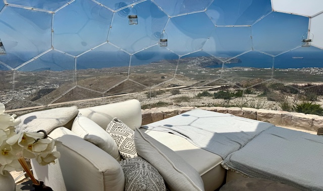 Panoramic island and sea view from inside a dome made of hexagonal glass pieces with a cozy bed set up at Santorini Sky in Pyrgos.