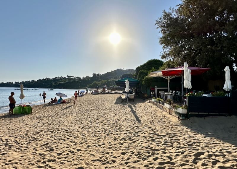 The sun hangs low in the sky over a golden beach with a taverna and water sports rental