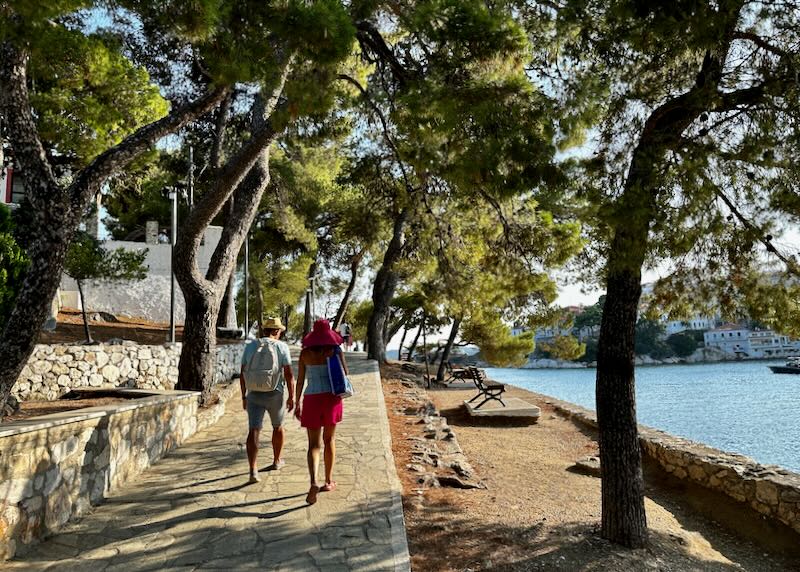 A couple walk along a stone path shaded by pine trees near the sea.