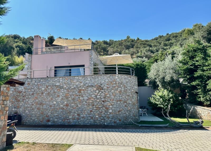 View looking up at a Greek villa made of stone, set on a pine-covered hillside