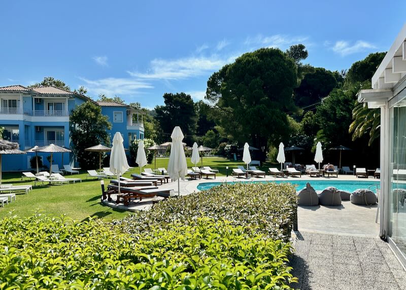 Hotel swimming pool in front of a light blue villa amid lush green landscaping.