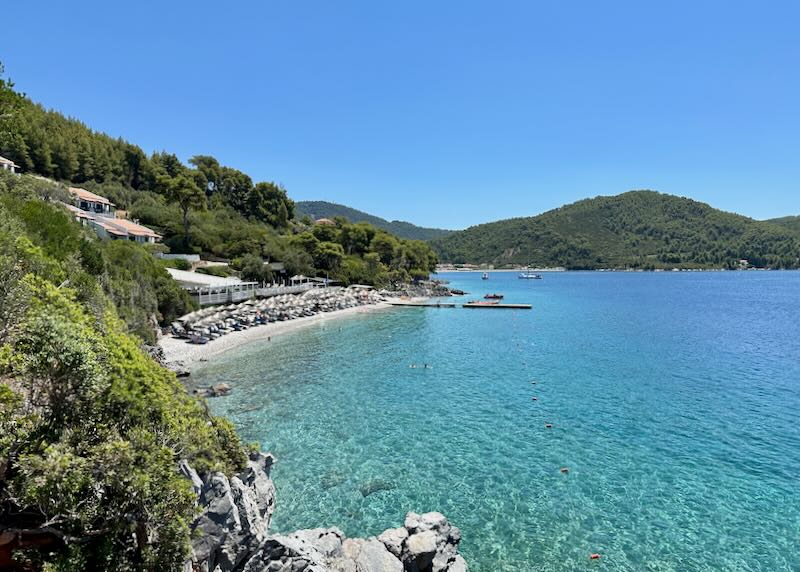 View from afar of a white beach on a pine covered island in Greece, lined with sun beds.