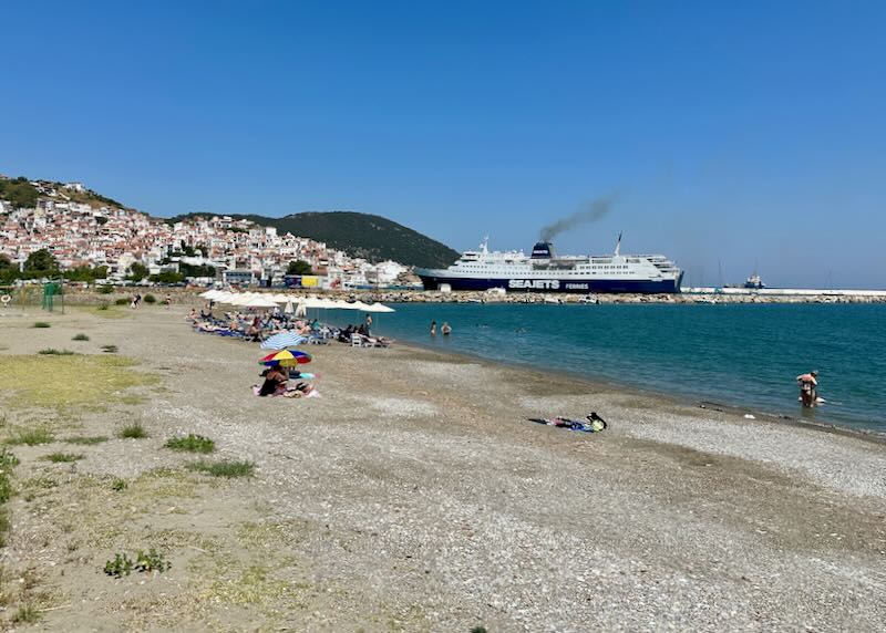 Small sandy beach near a town in Greece, with a ferry boat in the adjacent harbor