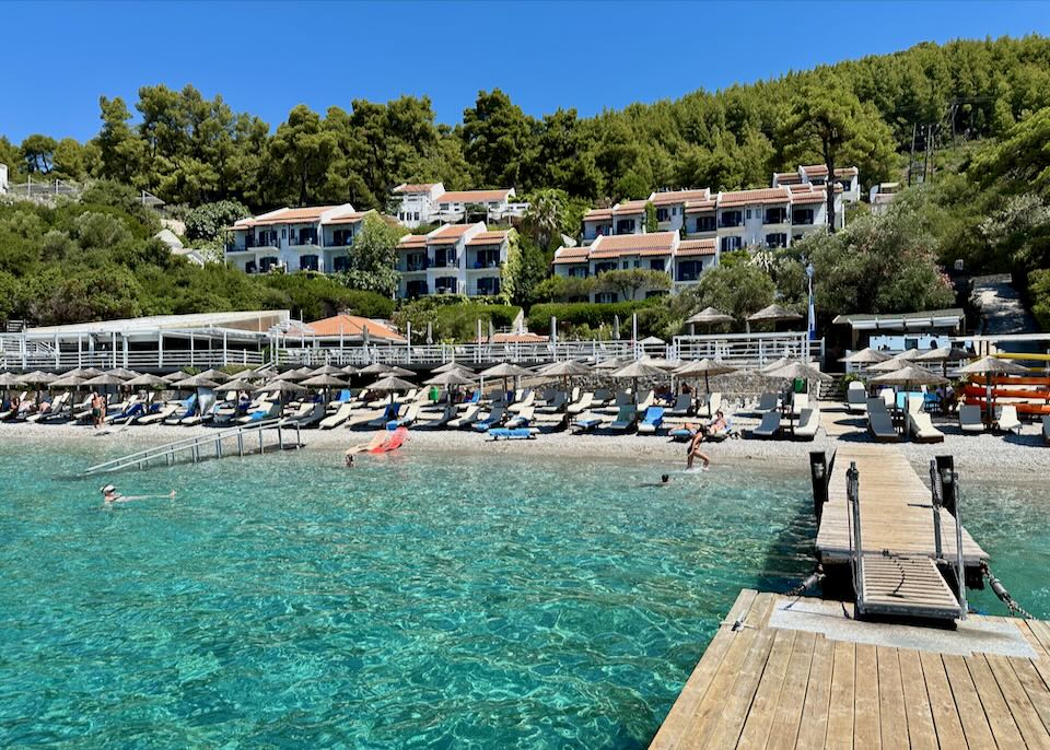 View from a dock over the water to a mid-sized hotel behind a white pebble beach.