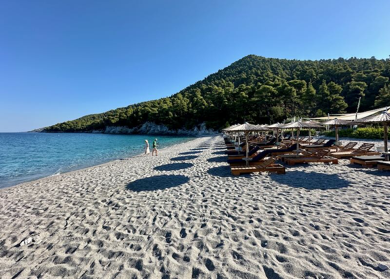 Pebbly beach lined with sun beds, their thatched umbrellas making circular shadows on the sand.