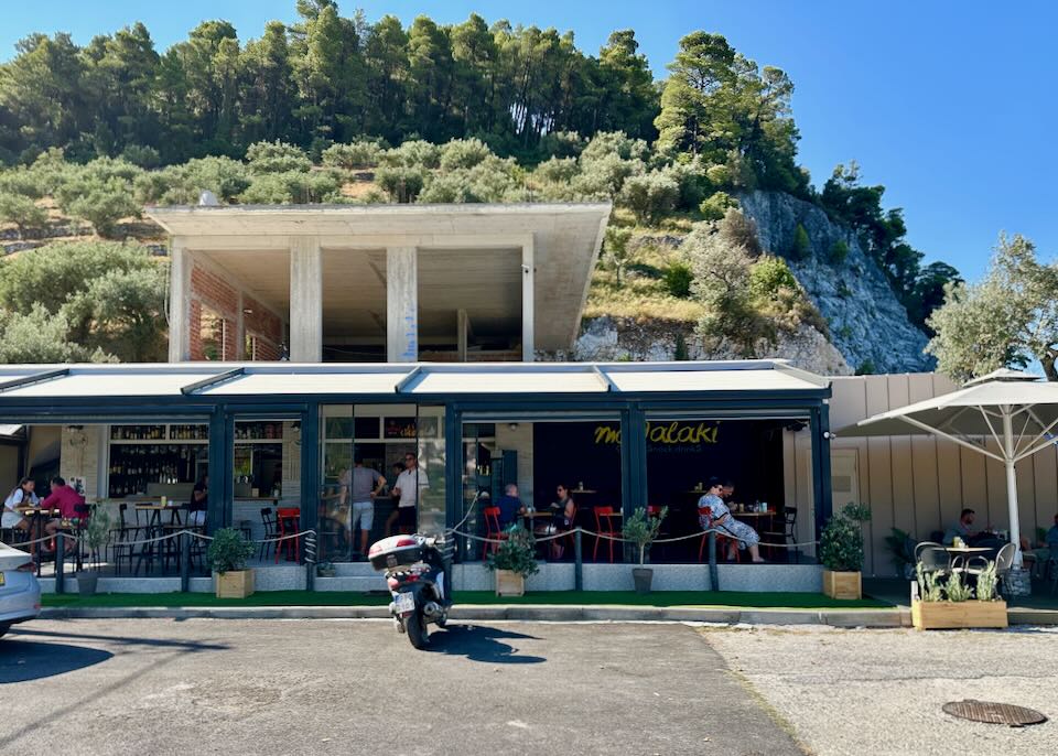 Motorbike parked outside of a small Greek taverna with open-air seating.