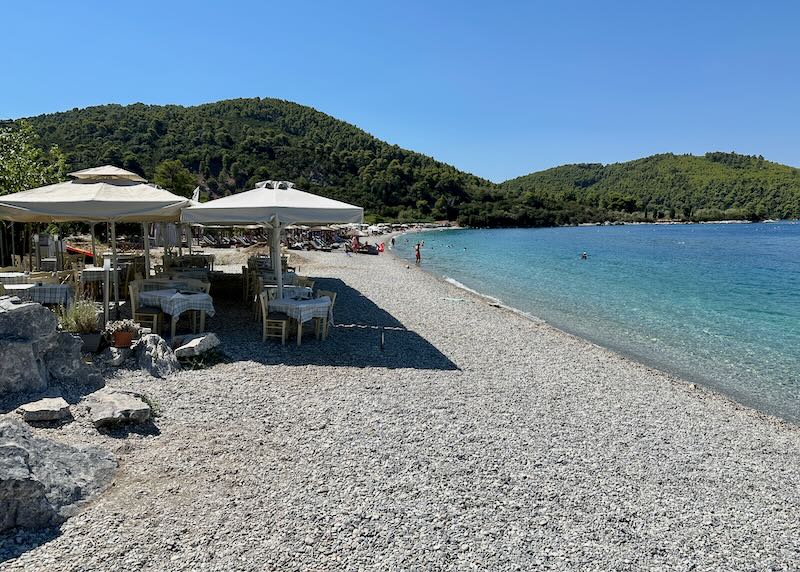 Cafe tables set in the sand and shaded by sun umbrellas on a beach in Greece.