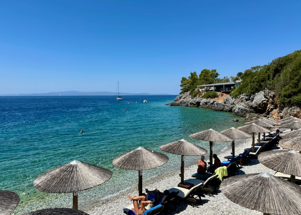 Thatched sun umbrellas line a pebbly, picturesque beach.