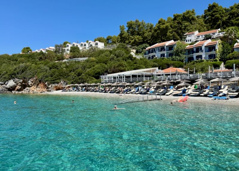 View from crystal blue water of a sandy beach with a large hotel rising up behind in the trees