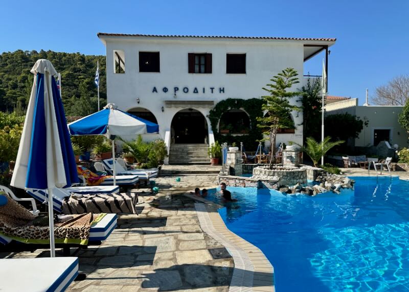 View from a stone swimming pool deck up to a traditional Greek hotel with arched porch colonnade