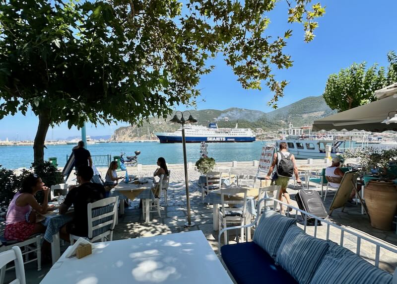 People dine at tree-shaded cafe tables next to a pretty Greek harbor.